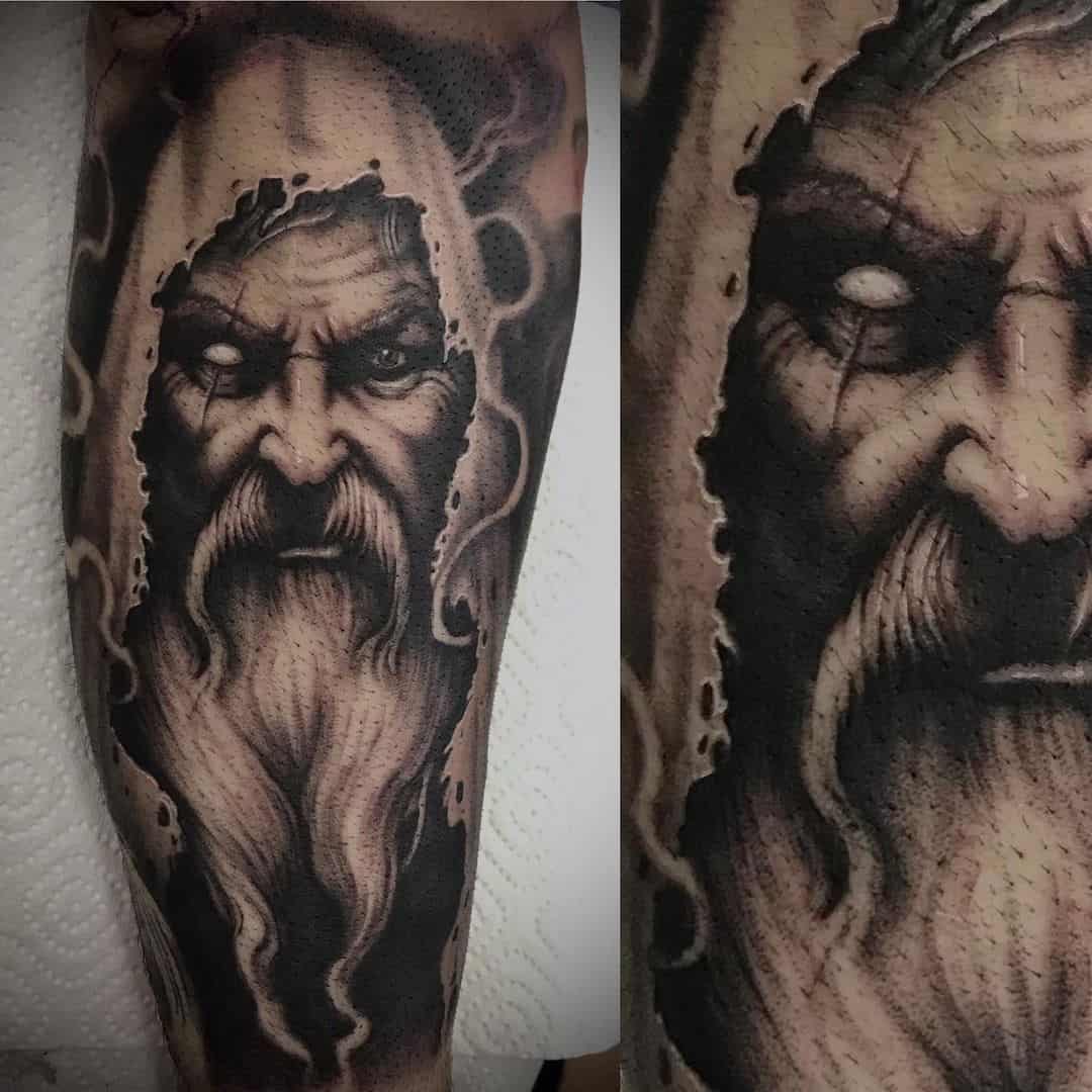 Black and grey realisme tattoo. Oude man met lange baard en kap op zijn hoofd. Linkeroog is wit met een litteken erdoor