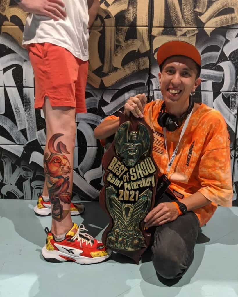 Tattoo artiest Kam Azat poseert met de eerste prijs van een conventie naast zijn klant. Zijn klant heeft een new school kleurentattoo van een konijn op het onderbeen.