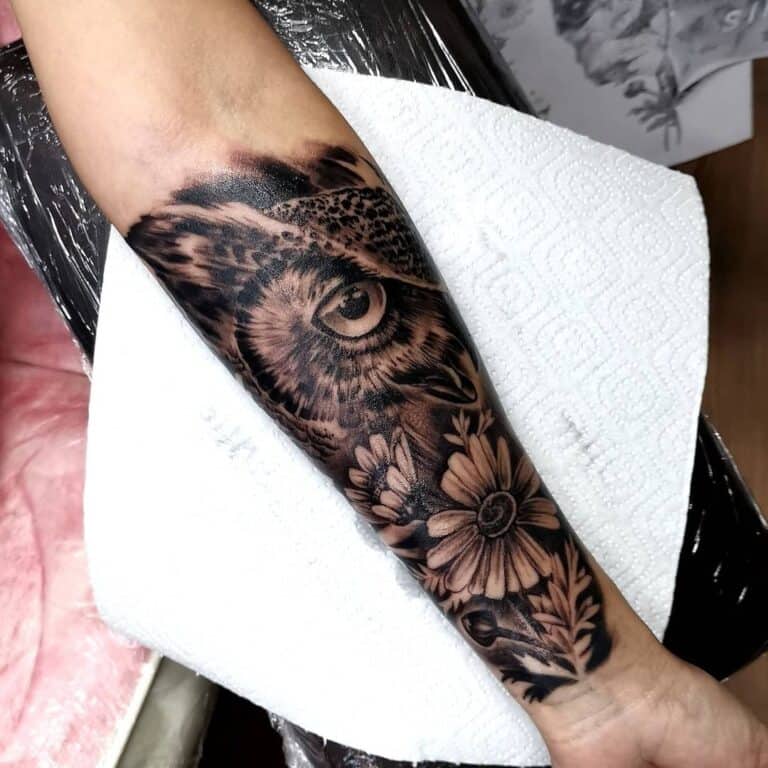 Black and grey realisme tattoo van een uil met bloemen op de onderarm.