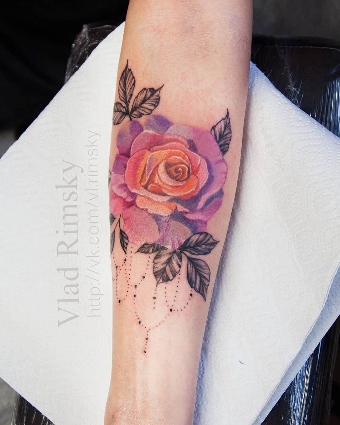 Realistische roos met oranje en paarse tinten met een achtergrond van black and grey bladeren en dotwork chain. Op een onderarm geplaatst bij Inksane tattoo en piercing.
