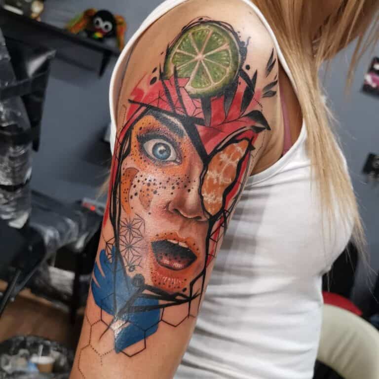 Surrealisme kleur tattoo op de bovenarm. Vrouwengezicht met fruit en geometrische vormen. Geplaatst bij Inskane tattoo en piercing.