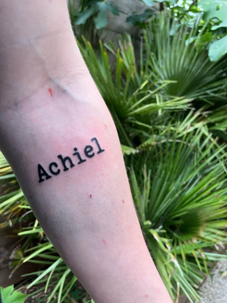 Tekst tattoo op de binnenkant van de onderarm. De naam Achiel in zwarte drukletters, in de stijl van een oude typemachine.